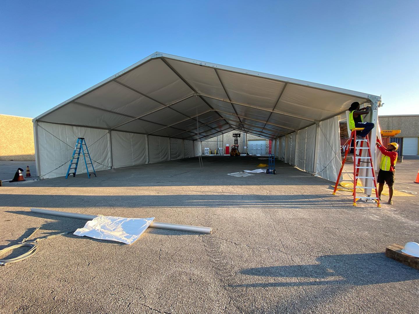Construction team members set up a large, open, white tent on pavement.