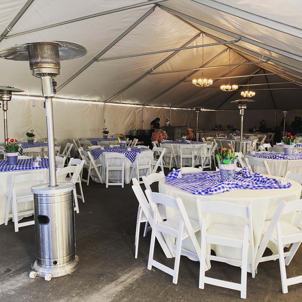 An outdoor tent containing white chairs around circular tables, ceiling chandeliers, and space heaters.