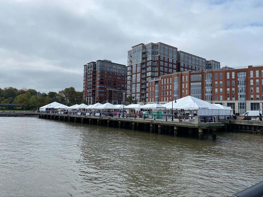 A pier on the water that contains several white tents for an event with tall buildings in the background.