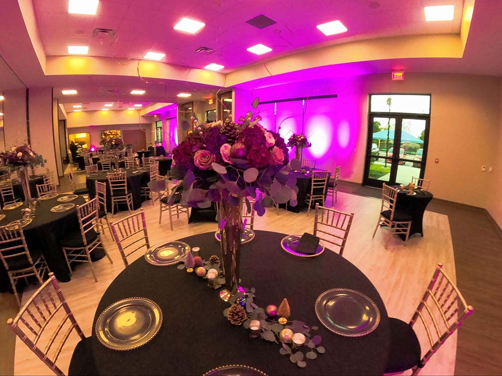 Decorated tables with black tablecloths, clear plates, a purple flower centerpiece, and gold chairs with a bright purple LED light in the background