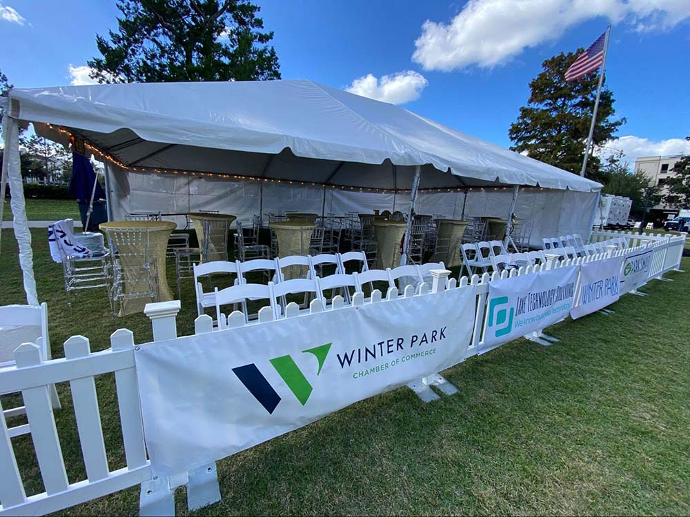 An outdoor tent with white folding chairs, a white fence border, and taller tables with clear tall chairs.